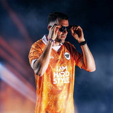 Brennan Heart wearing sunglasses and the orange "I AM HARDSTYLE" football shirt by Brennan Heart x COPA in a smoky, dramatic setting.