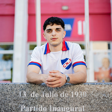 Man wearing a retro Nacional football shirt with red and blue trim, standing behind a stone monument at a stadium entrance.