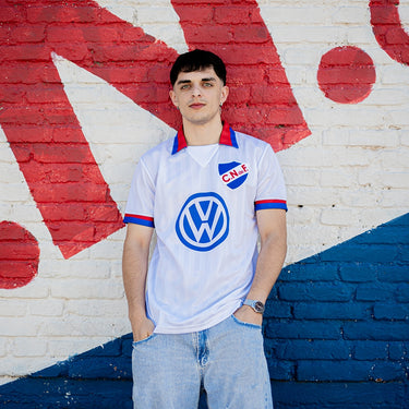 Man wearing a retro Nacional football shirt with Volkswagen sponsor, standing against a red, white, and blue mural wall.
