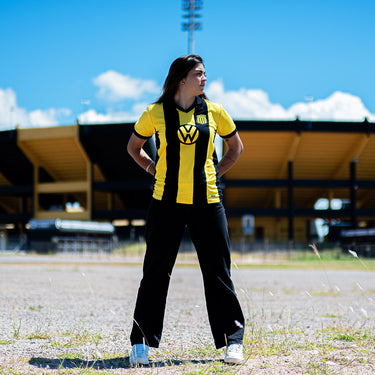 Woman wearing a retro Peñarol football shirt with Volkswagen sponsor, standing outside a stadium under a blue sky.

