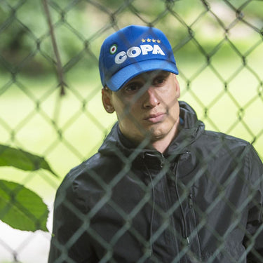 Man wearing a blue COPA trucker cap standing behind a chain-link fence